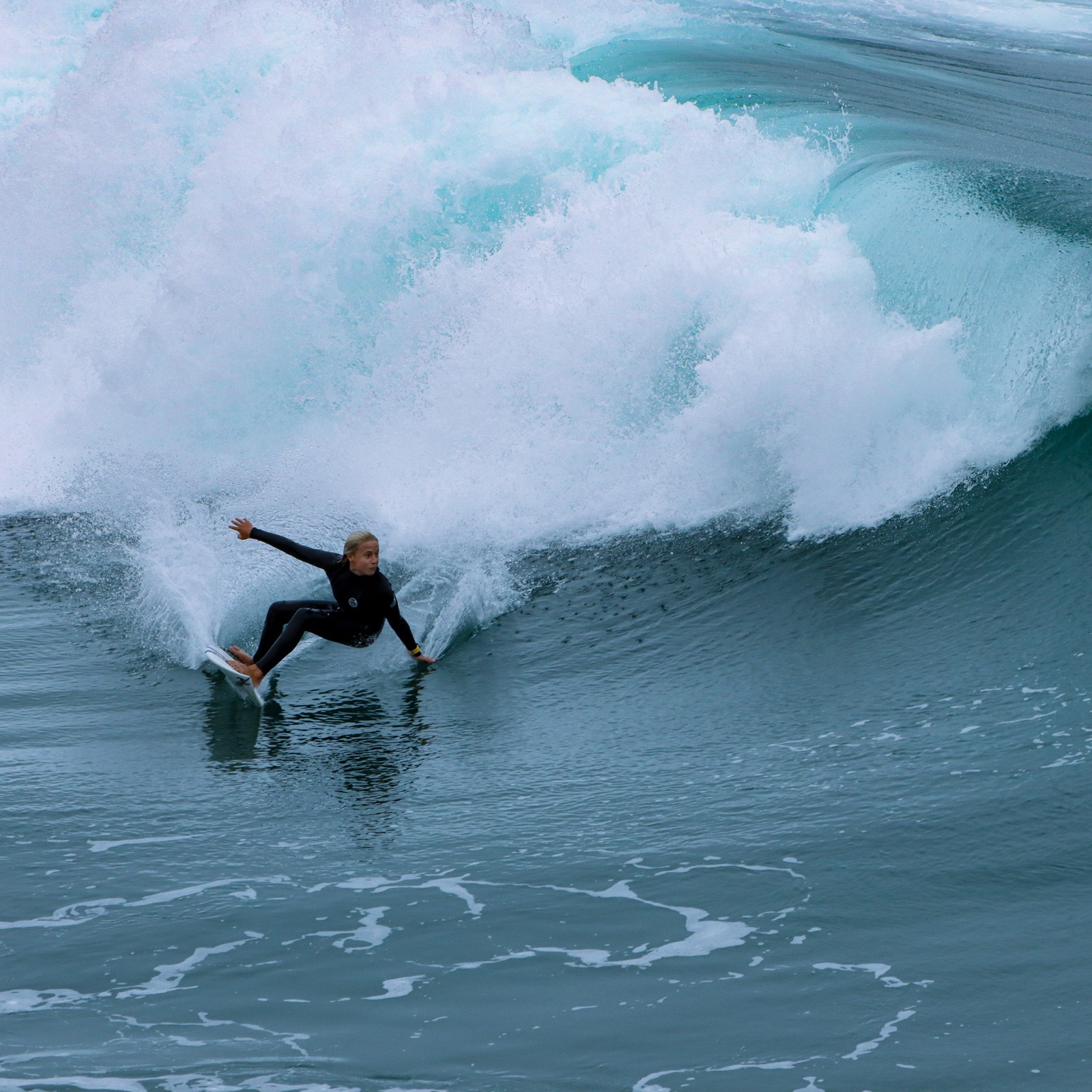 A surfer in a black wetsuit riding low along the face of a large breaking wave. Surf photography by Alice Njambi.